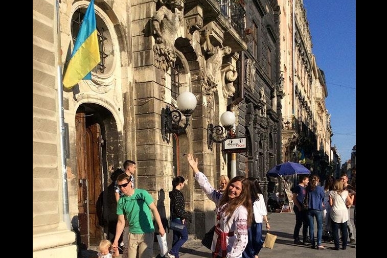 [ai] A woman with long brown hair poses with a smile in front of a historical building featuring a Ukrainian flag. Other people walk past, engaged in various activities, in a lively urban setting.